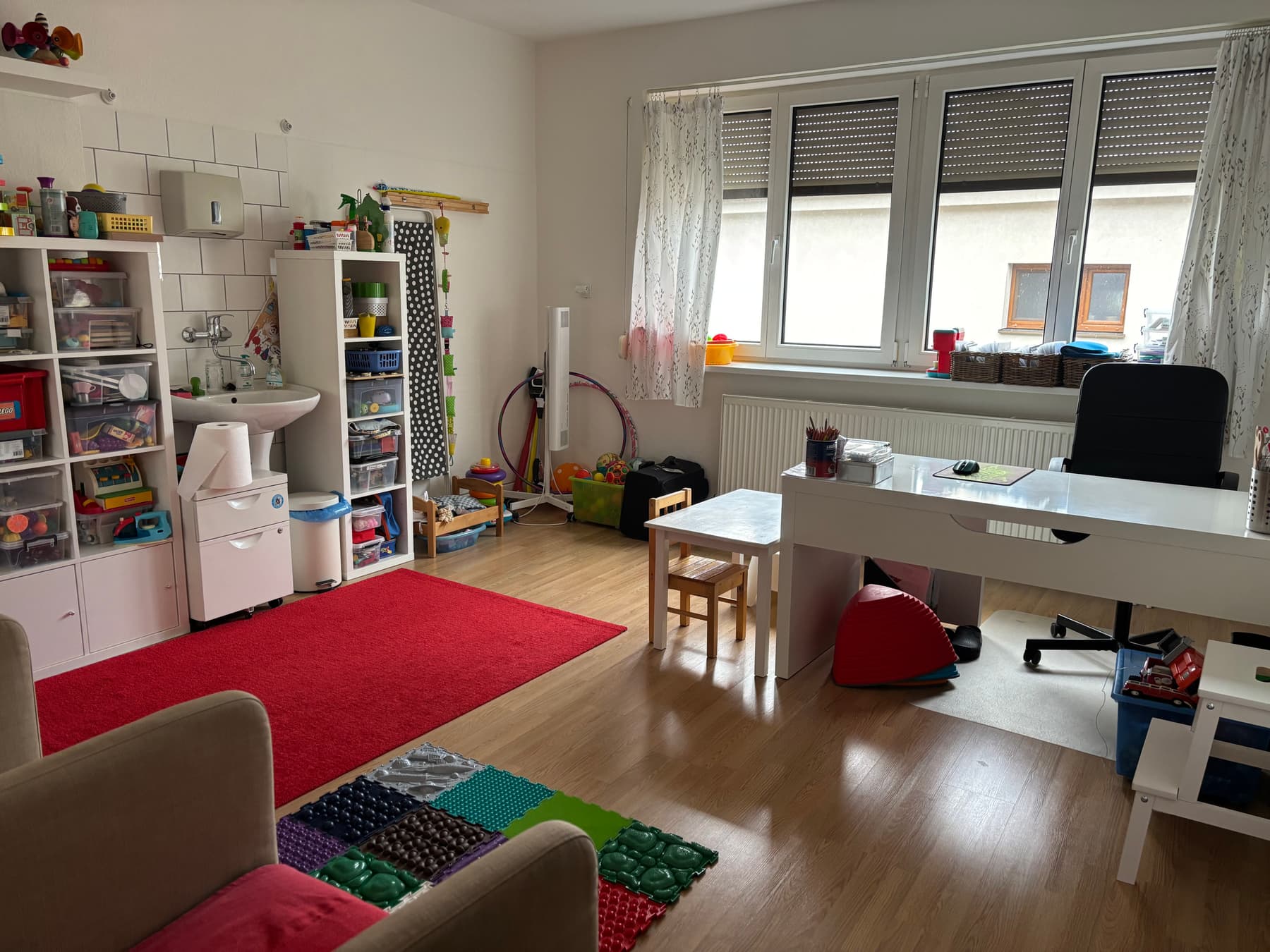 Terapeutická miestnosť ambulancie: regály s hračkami, červený koberec, senzorická podložka, detský stolík a stoličky pri okne.